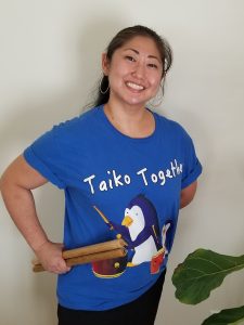 Our Team 5 Jana wearing a blue T-shirt with the Taiko Together parent-child penguins design. She is holding a pair of drum sticks in her hand.